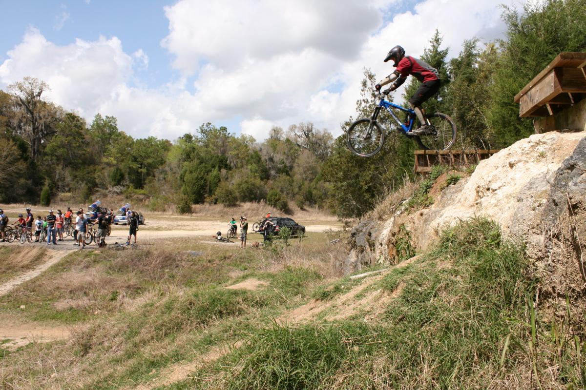 A mountain biker in a red shirt and helmet jumps off a ramp into the air, surrounded by spectators and a natural landscape with trees and clouds in the background. Santos mountain bike trail.