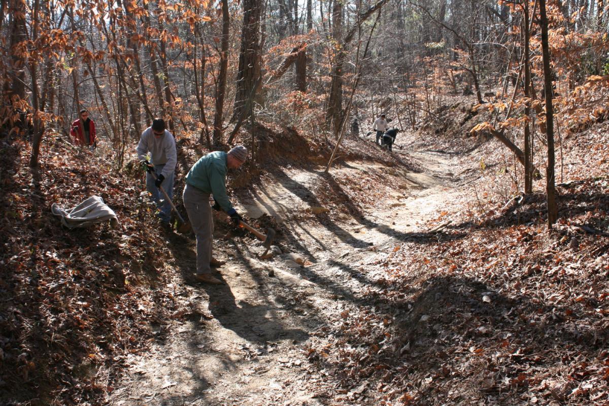 A group of volunteers working to clear a hiking trail in a wooded area during autumn. Fallen leaves cover the ground, and the sun shines through the trees. One individual is digging with a tool, while others are gathering or assisting nearby. Sope Creek mountain bike trail.