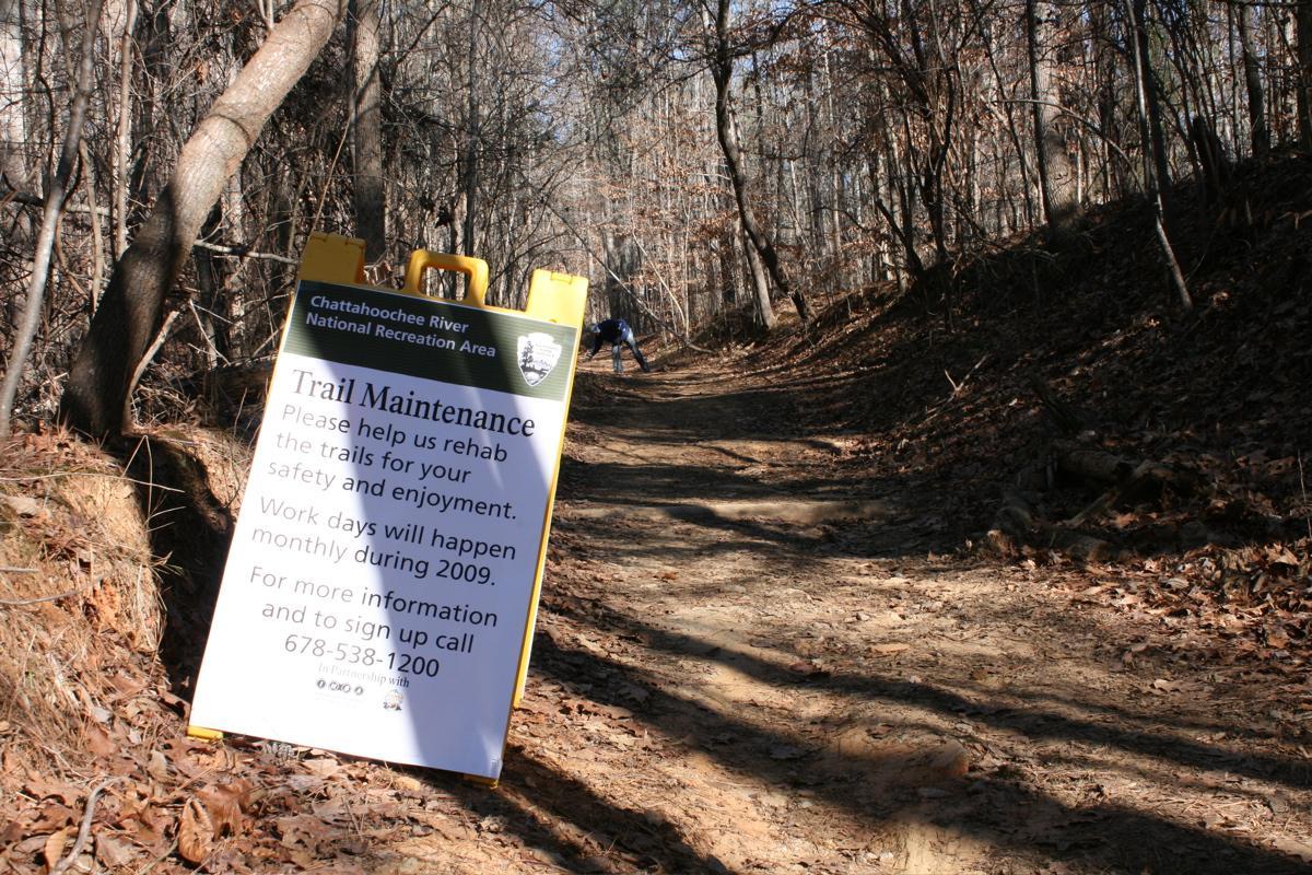 A trail sign at Chattahoochee River National Recreation Area announcing trail maintenance. The sign requests assistance for rehabilitating the trails for safety and enjoyment, with details about monthly workdays and a contact number for more information. In the background, a dirt trail is visible, bordered by trees and fallen leaves. Sope Creek mountain bike trail.