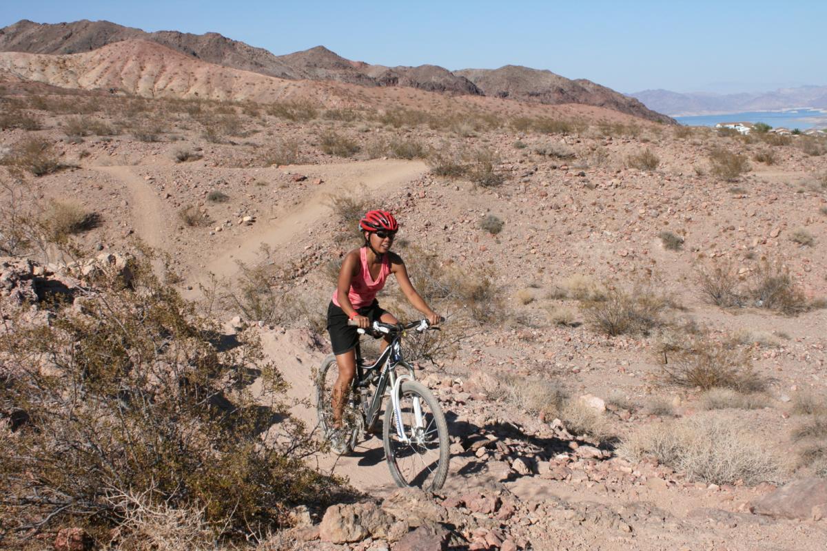 A person riding a mountain bike on a rocky trail in a desert landscape. The cyclist is wearing a red helmet and a pink sleeveless top, with dry vegetation and hills in the background. Clear blue skies and distant mountains are visible in the scenery. Bootleg Canyon mountain bike trail.