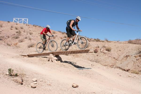 Two mountain bikers are seen riding over a wooden bridge on a dirt trail in a desert landscape. One biker in a red shirt is behind the other, who is wearing a black shirt and is airborne as he jumps off the bridge. The background features sandy terrain and sparse vegetation under a clear blue sky. Bootleg Canyon mountain bike trail.