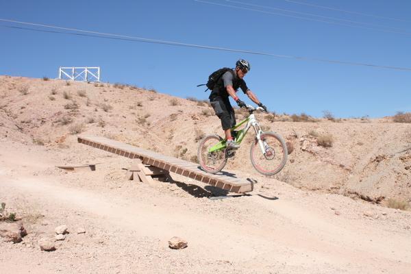 A mountain biker jumping off a wooden ramp on a dirt trail, surrounded by rocky terrain under a clear blue sky. The rider is in motion, wearing a helmet and protective gear, showcasing an active outdoor adventure. Bootleg Canyon mountain bike trail.