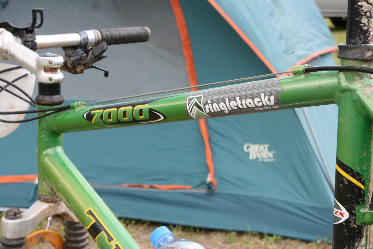 A close-up image of a green mountain bike frame featuring logos and markings, including "7000" and "singletracks." In the background, a camping tent with blue fabric and orange trim is visible, along with a partially visible water bottle on the ground. Santos mountain bike trail.