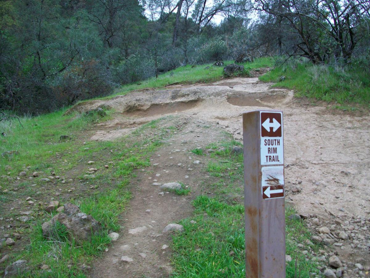 A dirt path leading into a grassy area, with a signpost indicating the direction of the South Rim Trail. The surrounding landscape features trees and rocky terrain. Upper Bidwell Park mountain bike trail.