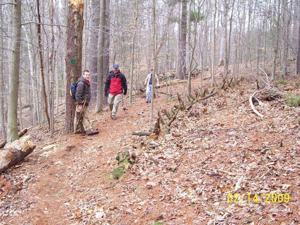 Three individuals are walking along a dirt trail in a wooded area. The ground is covered with pine needles and fallen leaves, and several trees are visible in the background. The scene captures a cool, overcast day in a nature setting. North Bend State Park mountain bike trail.