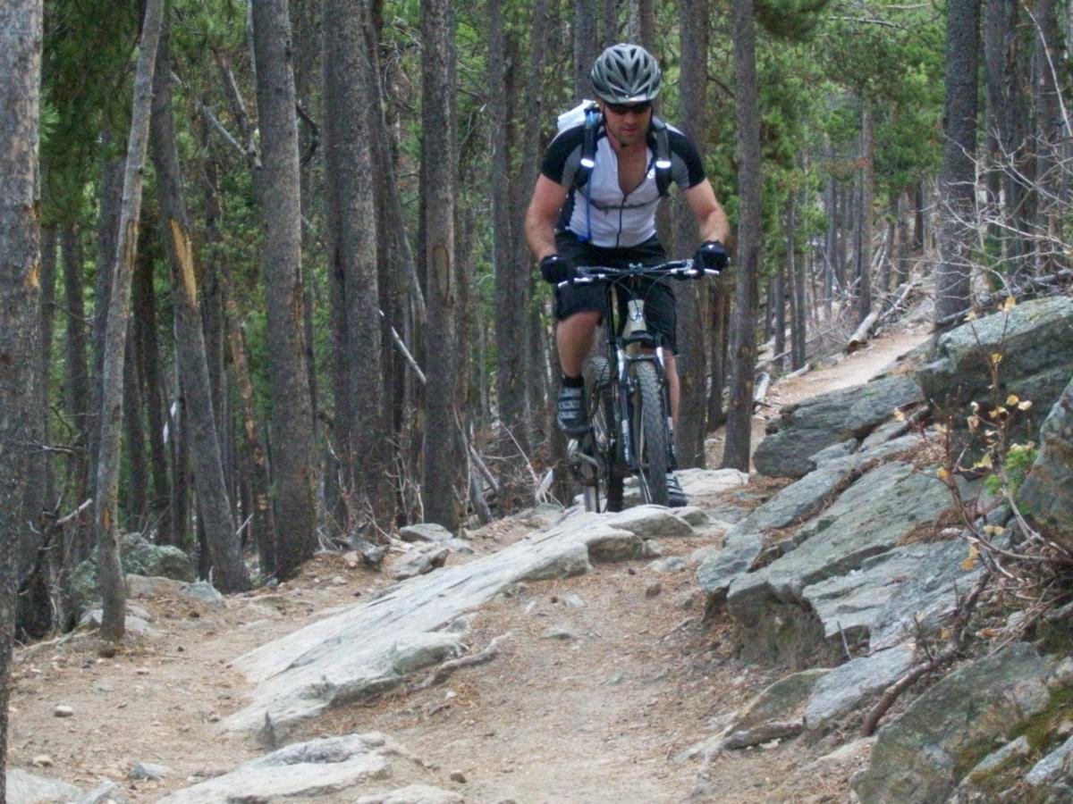 A mountain biker navigating a rocky trail through a dense forest of pine trees, wearing a helmet and cycling gear. Bergen Peak mountain bike trail.