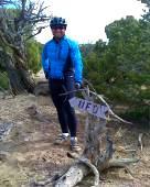 A cyclist wearing a blue jacket and helmet stands next to a hand-painted sign pointing towards "UFO," surrounded by a natural landscape with trees and a rocky path. Alien Run Trail mountain bike trail.