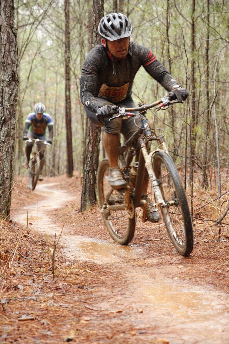 Two mountain bikers riding on a muddy dirt trail through a forest. The first rider is in the foreground, wearing a helmet and a muddy jersey, while the second rider can be seen behind on a similar trail. Pine trees surround the path, and the ground is covered with pine needles and puddles from recent rain. Blankets Creek mountain bike trail.