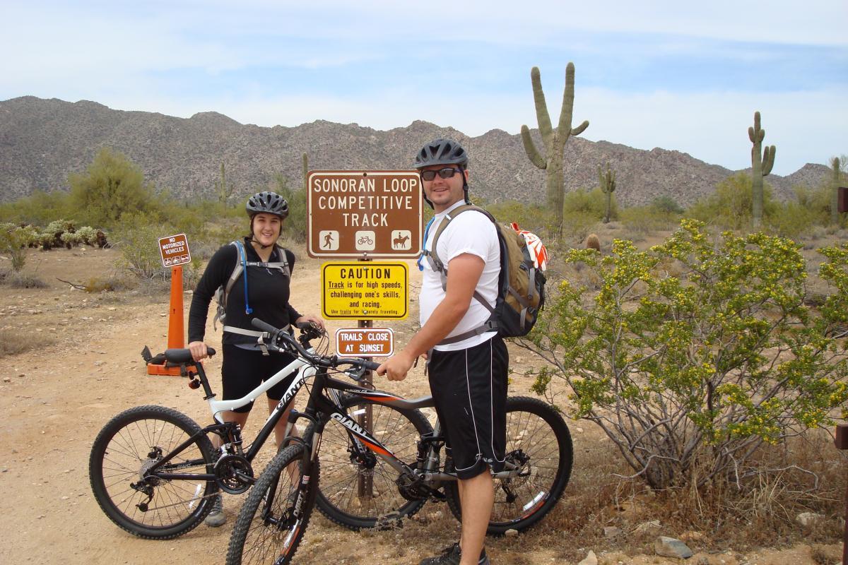 Two mountain bikers stand next to their bikes at the Sonoran Loop Competitive Track. A sign in the background displays warnings and guidelines for the trail, surrounded by a desert landscape with cacti and mountains. The riders wear helmets and are dressed in outdoor gear, ready for their cycling adventure. White Tanks Competitive Track mountain bike trail.