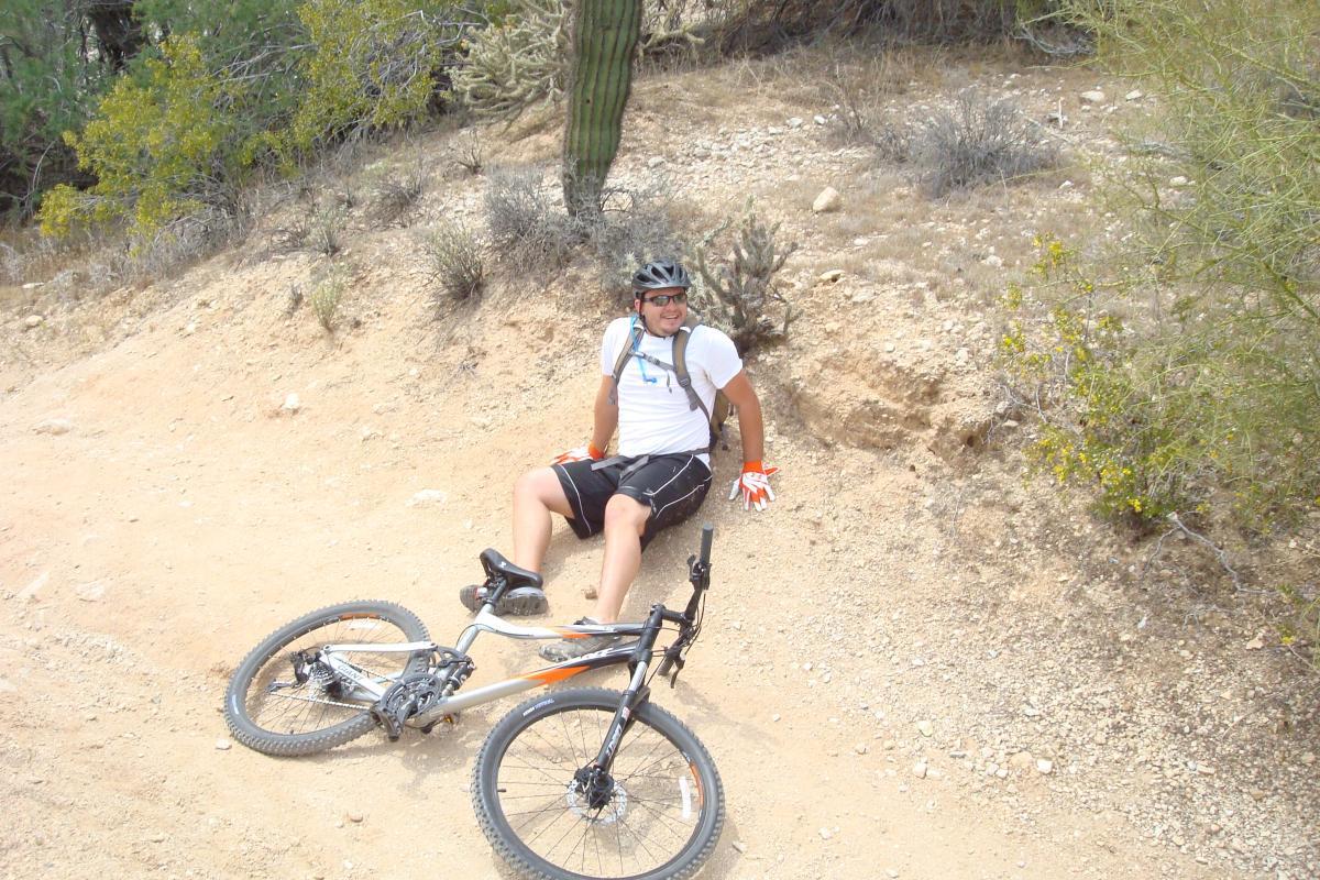 A person sitting on a dirt trail next to a mountain bike, wearing a helmet, gloves, and casual sports attire. Surrounding vegetation includes desert plants and cacti, with a rocky and sandy terrain in the background. The individual appears relaxed after a ride. White Tanks Competitive Track mountain bike trail.