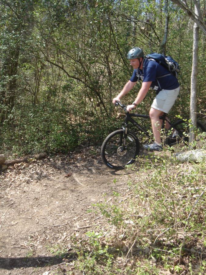 A person riding a mountain bike along a dirt trail, surrounded by greenery and trees. The cyclist is wearing a helmet and a backpack, and the path appears to be slightly uneven, indicating an adventurous biking experience. Lock 4 mountain bike trail.