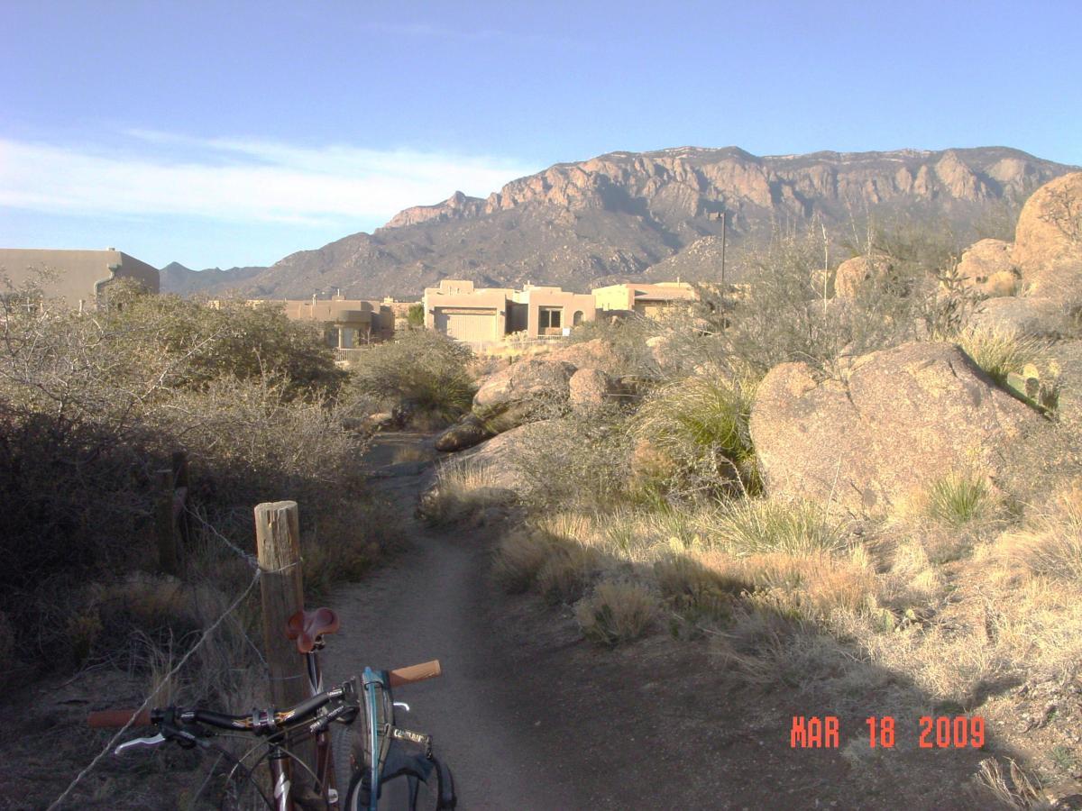 A dirt path leading through desert vegetation, with a bicycle leaning against a wooden post. In the background, adobe-style buildings and rugged mountains are visible under a blue sky. The image is dated March 18, 2009. Sandia Mountains Foothill Trail mountain bike trail.