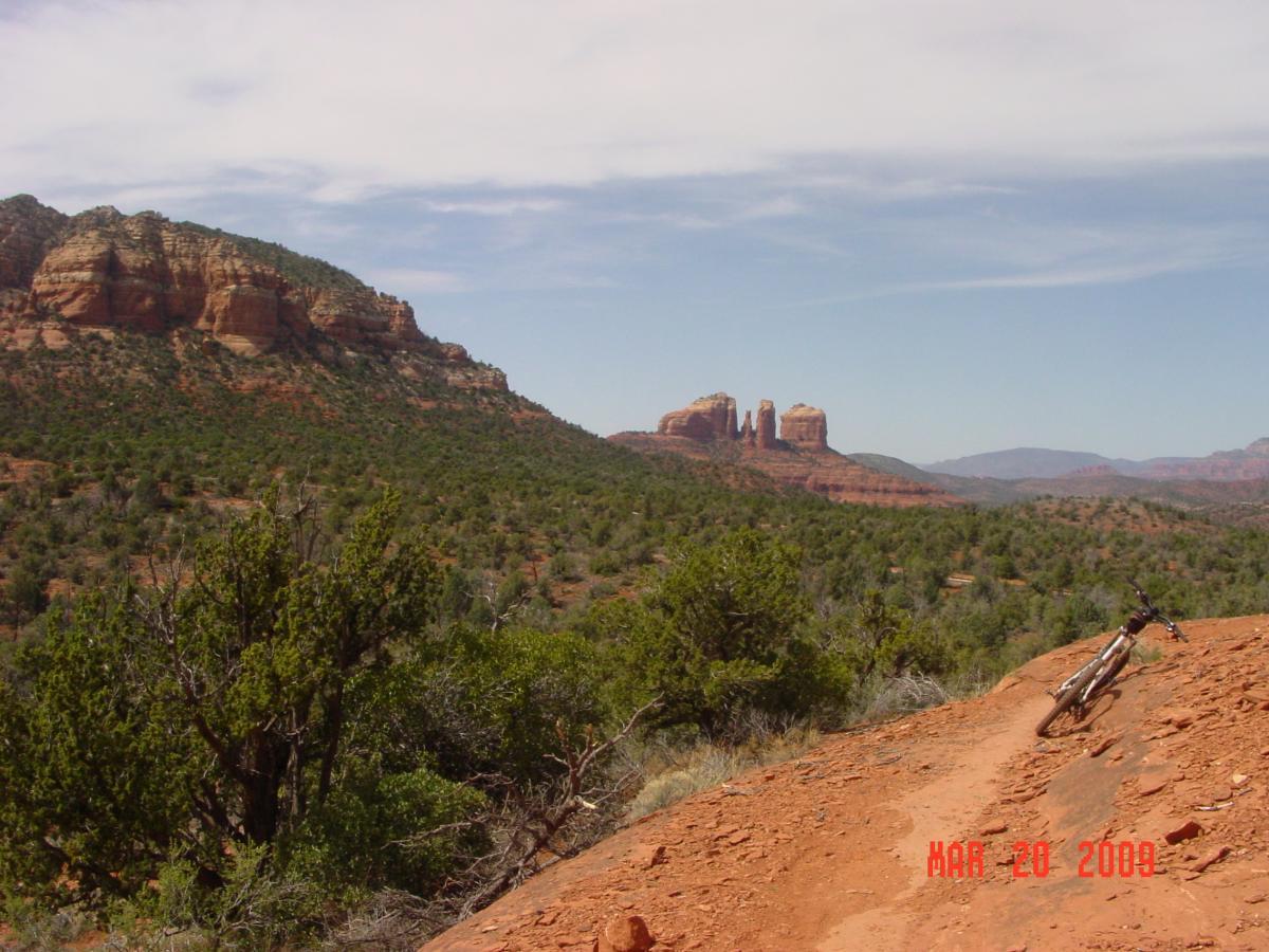 A scenic view of a mountainous landscape featuring red rock formations and lush green foliage. A dirt trail winds through the foreground, with a mountain bike leaning against the trail's edge. In the background, distinctive rock formations rise against a clear blue sky, suggesting a popular outdoor recreation area. Bell Rock Trailway mountain bike trail.