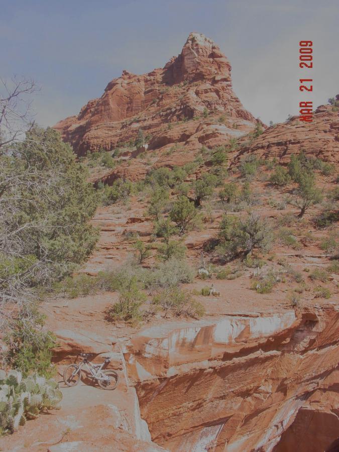 A mountain bike parked on a rocky trail surrounded by red rock formations and sparse vegetation, with a towering cliff in the background under a clear sky. The date "APR 21 2009" is displayed in the corner. North Urban Trail System - East mountain bike trail.