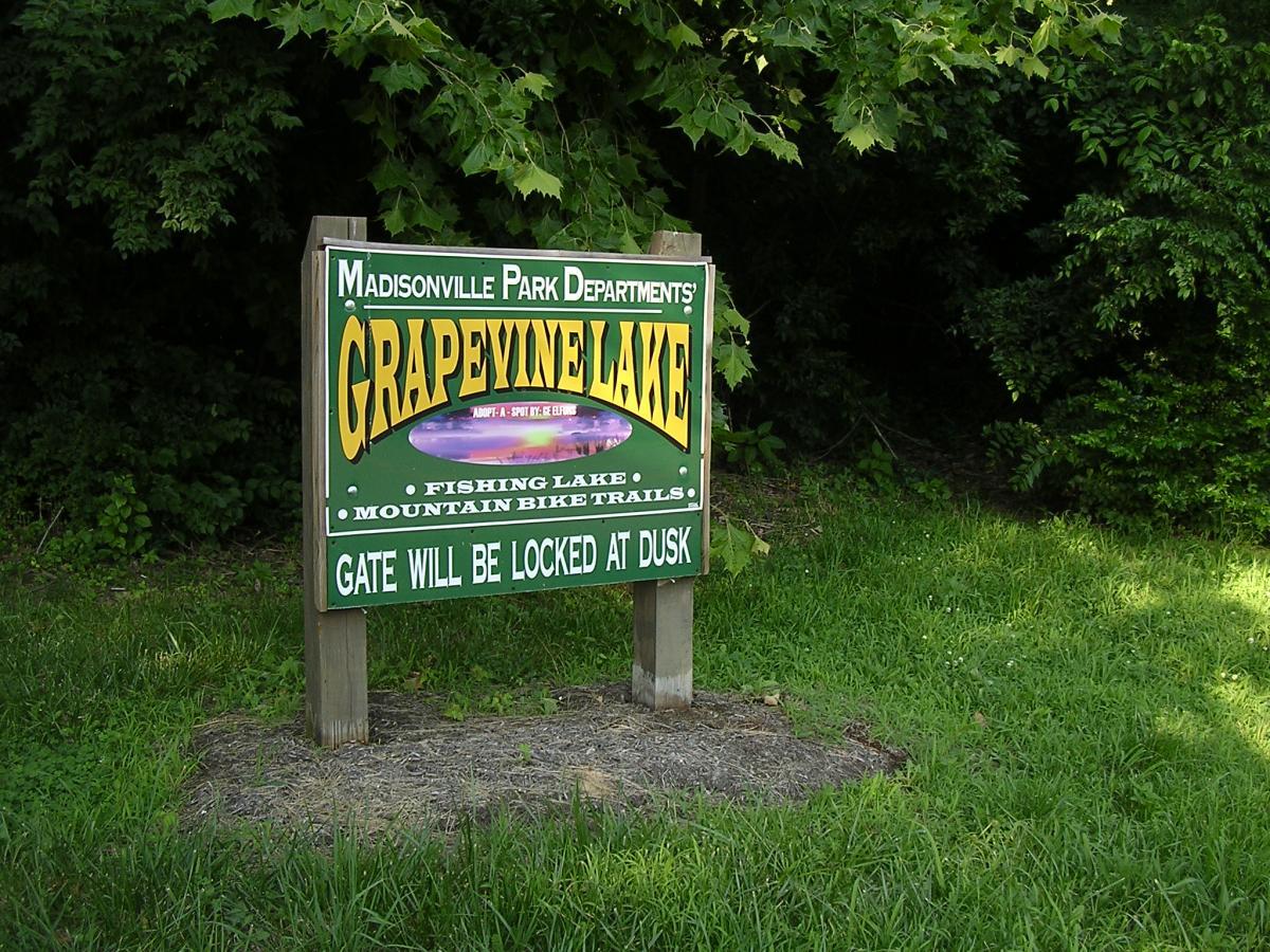 Sign for Grapevine Lake, featuring information about fishing and mountain bike trails, with a note that the gate will be locked at dusk. The sign is surrounded by greenery and grass. Grapevine Lake  A&amp;b mountain bike trail.