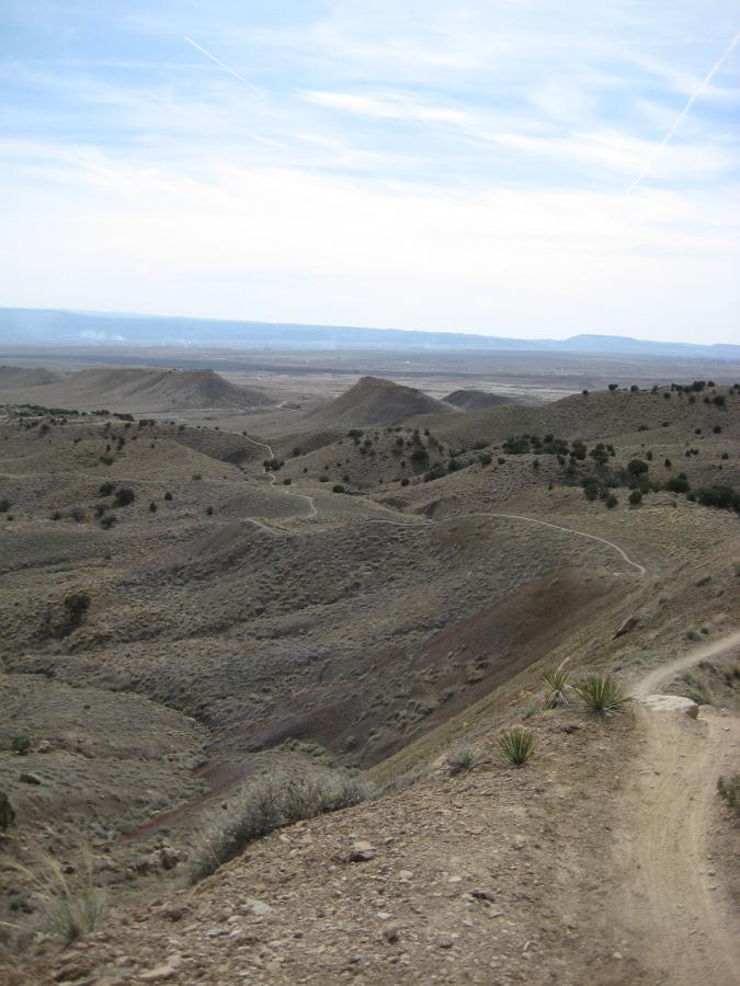 A panoramic view of rugged, undulating hills in a vast landscape. The scene features sparse vegetation, including small shrubs and grasses, under a partly cloudy sky. A dirt path winds through the terrain, leading into the distance, showcasing the natural beauty of the area. Joe's Ridge mountain bike trail.