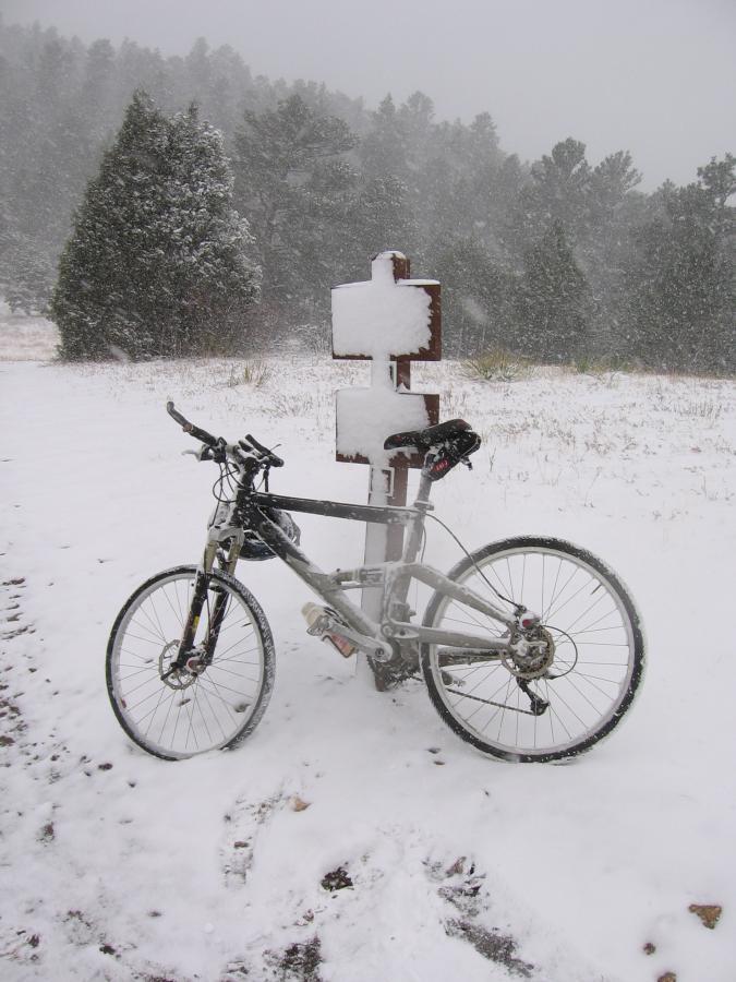 A mountain bike parked beside a snow-covered trail sign in a snowy landscape, with falling snowflakes and pine trees in the background. The ground is blanketed in snow, creating a winter scene. Centennial Cone Park mountain bike trail.