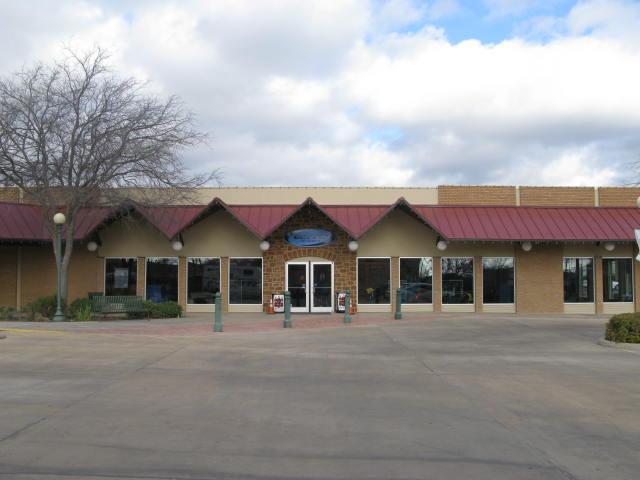 A building with a sloped red metal roof and stone accents, featuring large windows and a main entrance with double doors. The surrounding area includes a paved walkway and benches, with a few trees nearby. The sky is partly cloudy.