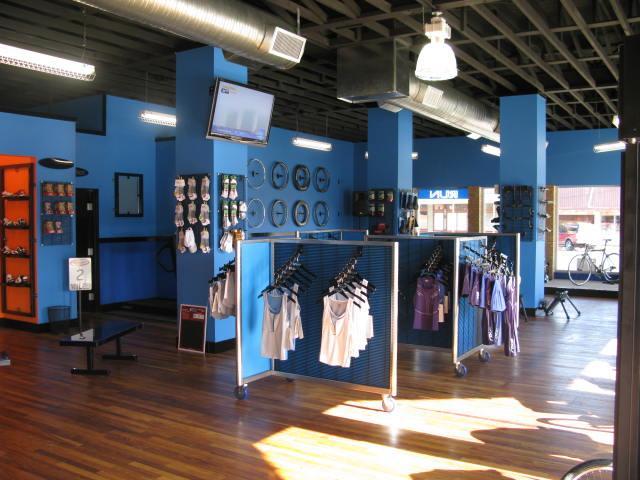 Interior of a retail store featuring blue walls, wooden flooring, and bright lighting. The space is organized with racks displaying athletic apparel and accessories, including tanks and shorts. A flat-screen TV is mounted on one wall, and there are shelves stocked with various products. Large windows allow natural light to illuminate the store.