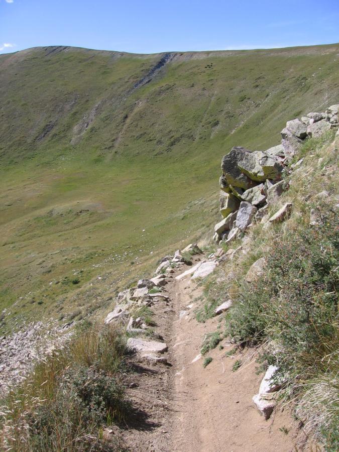 A narrow dirt path runs along the edge of a grassy hillside, bordered by scattered rocks and patches of vegetation. The path slopes downward, leading into a lush green valley in the background, under a clear blue sky. Canyon Creek Trail mountain bike trail.