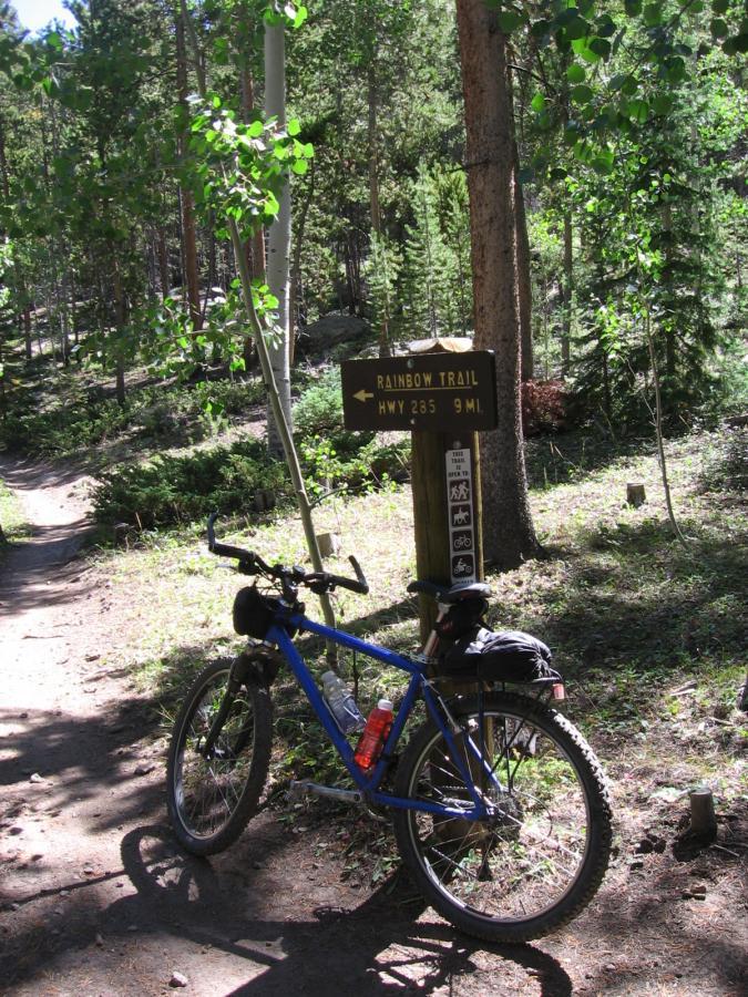 A blue mountain bike resting on the dirt trail beside a wooden sign indicating "Rainbow Trail" and directions to Highway 285, 9 miles away. The scene is surrounded by lush greenery and tall trees, showcasing a sunny outdoor environment. Rainbow Trail: Silver Creek to Hwy 285 mountain bike trail.