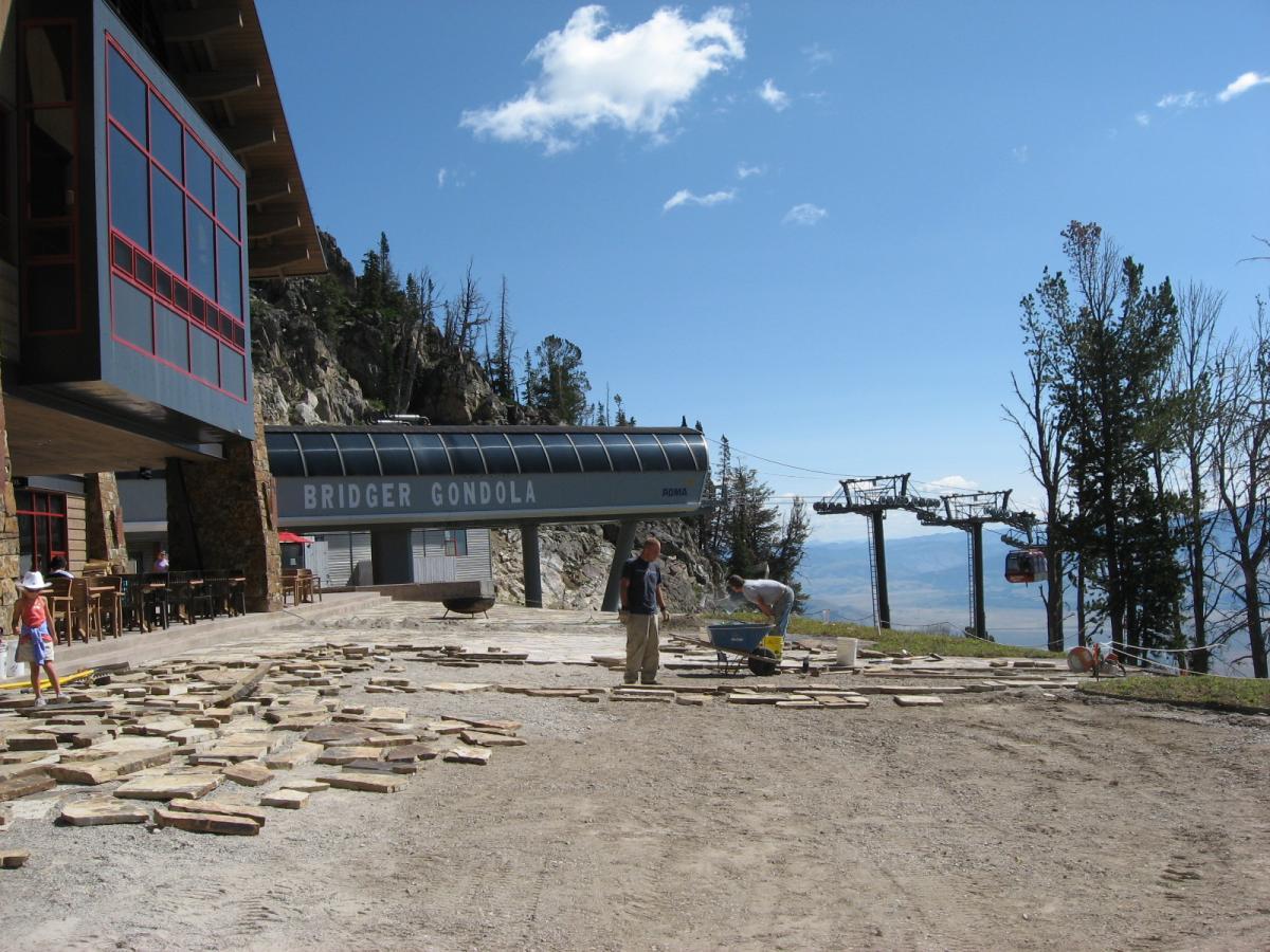 Construction site in front of the Bridger Gondola with workers laying stone tiles on a gravel surface. The gondola station is visible in the background alongside ski lift chairs, with a mountainous landscape under a clear blue sky. Jackson Hole Bike Park mountain bike trail.