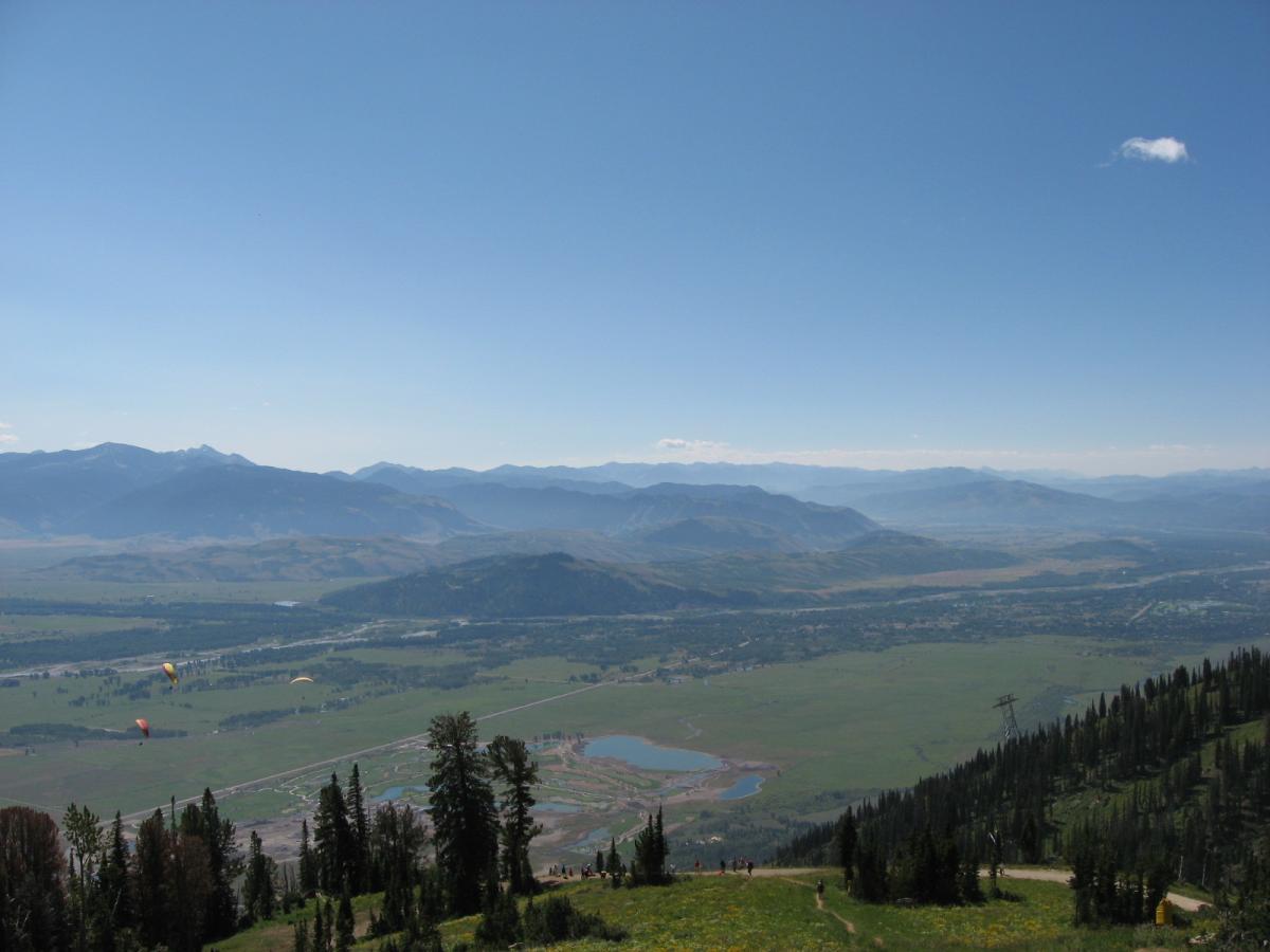 A panoramic view of a mountainous landscape under a clear blue sky. The foreground features green trees and a winding path, while the background displays a range of mountains fading into the distance. A few colorful paragliders can be seen soaring over the lush valleys and rivers below. Jackson Hole Bike Park mountain bike trail.
