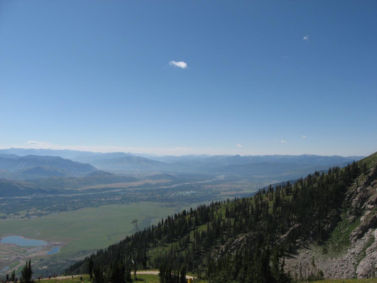 A panoramic view of a mountainous landscape under a clear blue sky, showcasing rolling hills and distant mountain ranges. The foreground features a steep, green slope with pine trees, while a valley below is dotted with patches of water and fields. Jackson Hole Bike Park mountain bike trail.