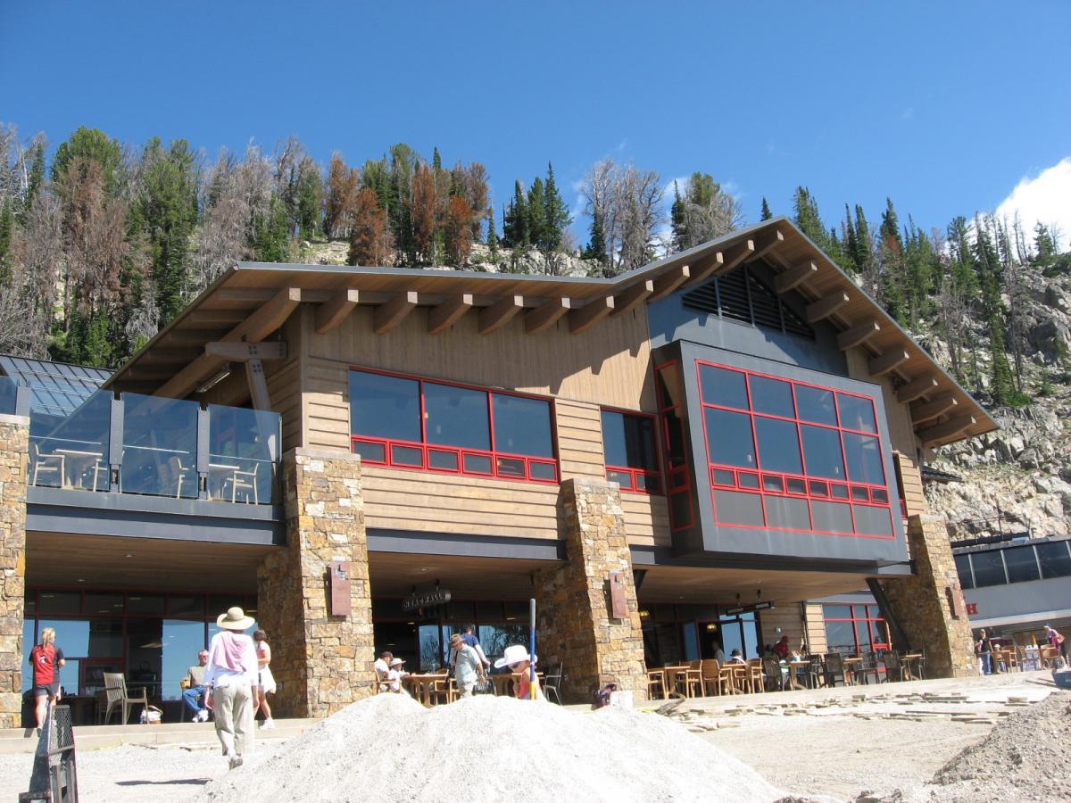 A modern wooden building with large red-framed windows and a stone foundation, set against a backdrop of green trees and a clear blue sky. The area in front features a gravel surface and scattered people enjoying the space, with some outdoor seating visible on a deck. Jackson Hole Bike Park mountain bike trail.