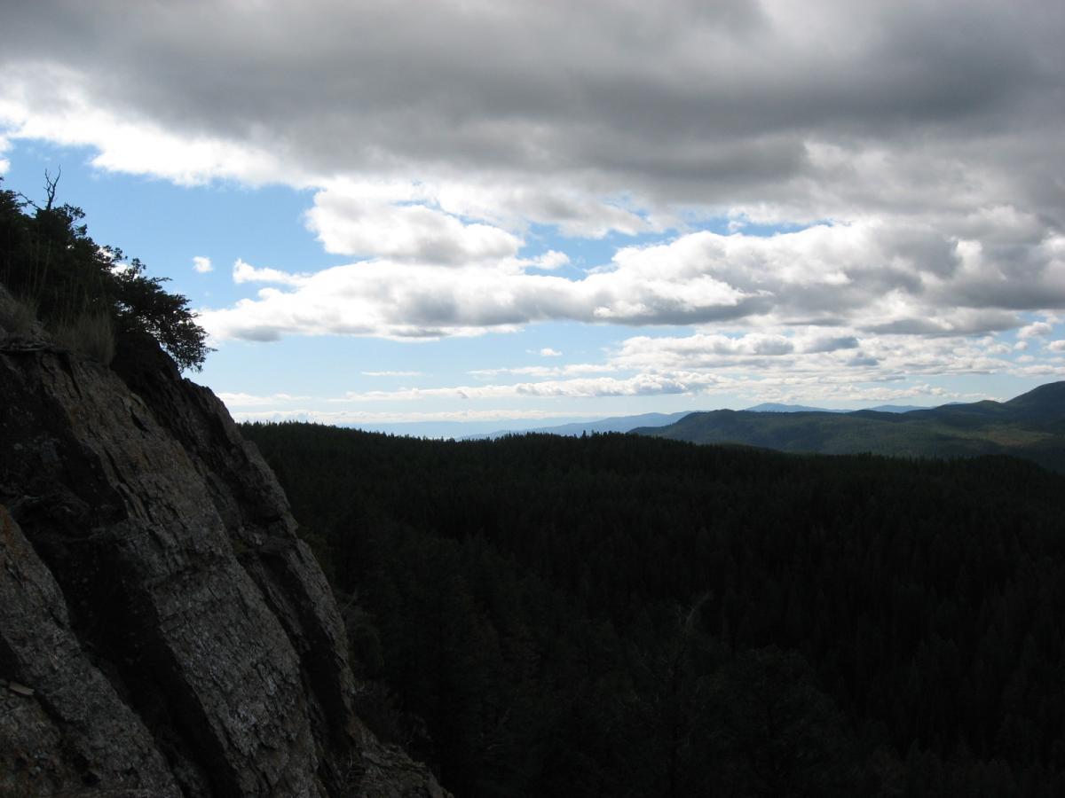 A panoramic view of a mountainous landscape showcasing a rocky cliff in the foreground, framed by lush green forests below and distant rolling hills. The sky is partly cloudy, with patches of blue peeking through the clouds. Tally Lake Overlook mountain bike trail.