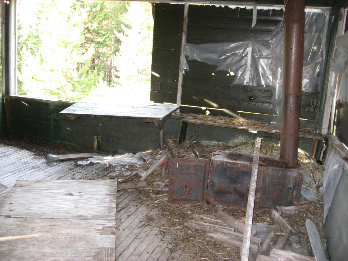 Interior of an abandoned building with peeling green walls, wooden floors covered in debris, and a rusted metal stove in the corner. Large windows reveal a view of trees outside, while one table is visible, partly covered in debris. Sunlight filters through the openings, highlighting the neglect. Johnson Peak/Trixie Pass mountain bike trail.