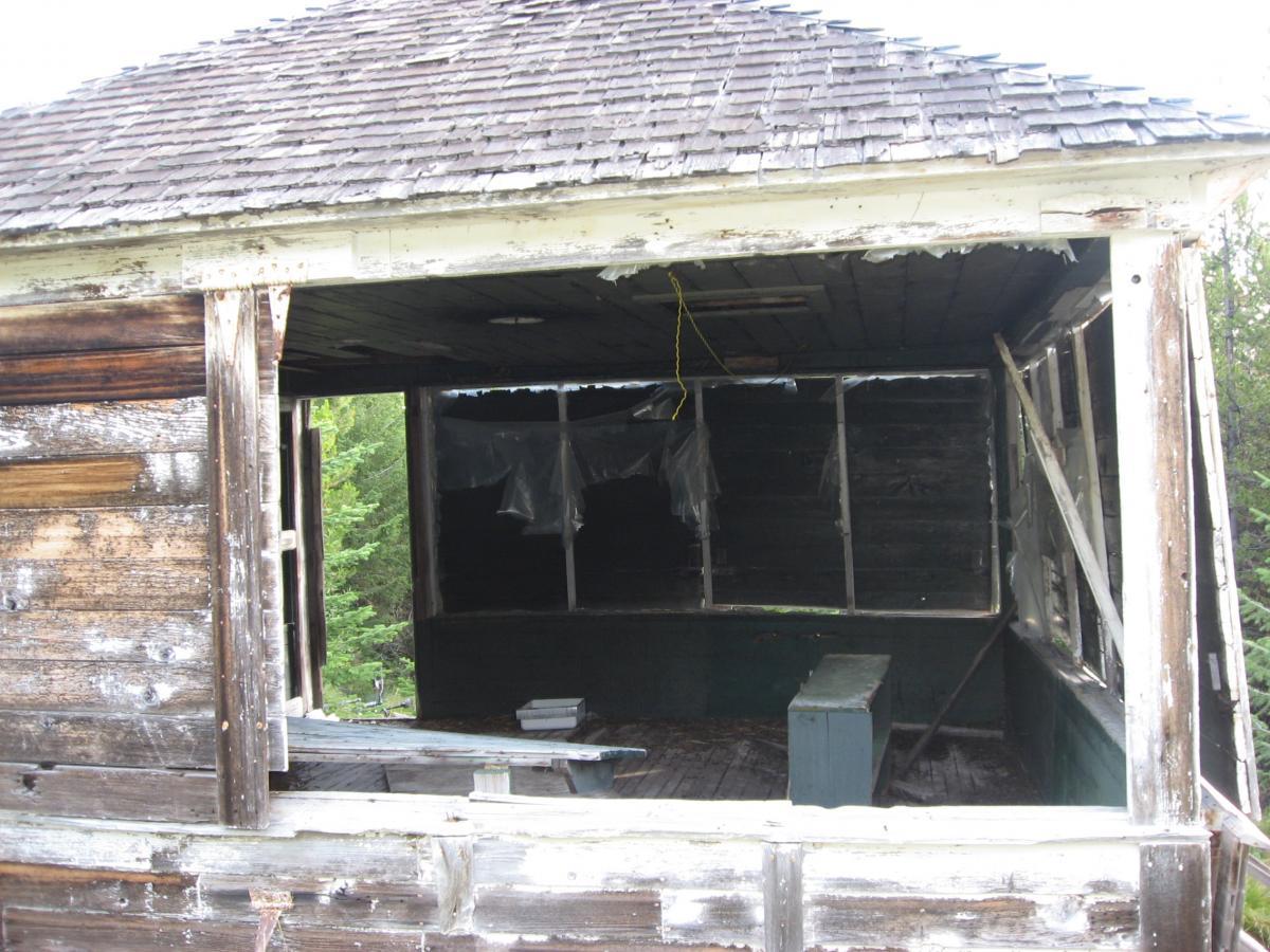 Alt text: A dilapidated wooden structure with a partially collapsed roof and open walls, revealing a dusty interior with a small table. The surrounding area features greenery, suggesting an overgrown environment. Johnson Peak/Trixie Pass mountain bike trail.