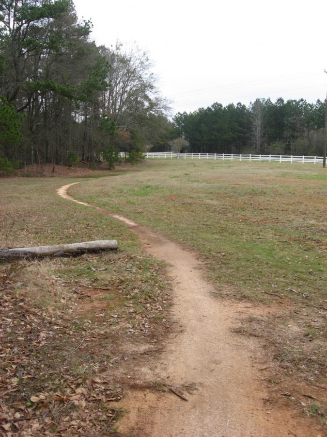 A winding dirt path leads through a grassy area, bordered by trees on the left and a white wooden fence on the right. The sky is overcast, hinting at a cool, serene atmosphere. Heritage Park mountain bike trail.