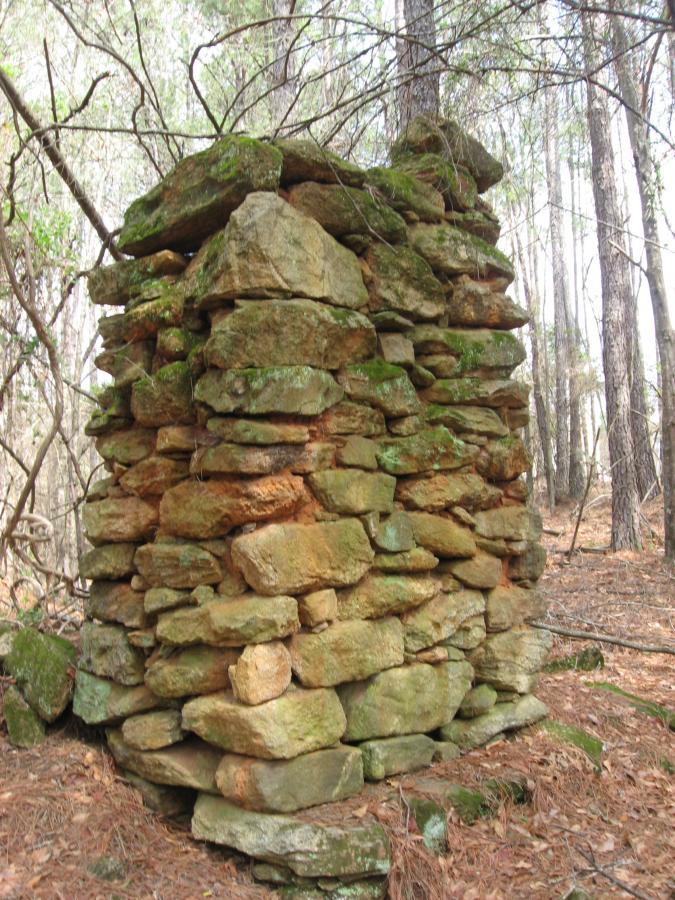 A weathered stone structure partially covered in moss, standing amidst a forest of tall trees. The stones are unevenly stacked, showing signs of age and natural wear, with fallen leaves and pine needles scattered on the ground around it. Heritage Park mountain bike trail.