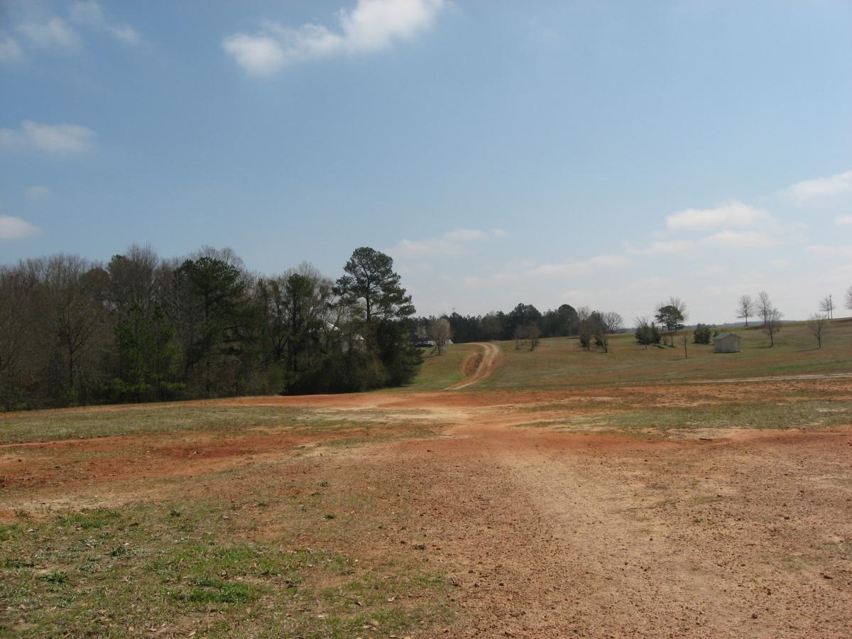 A wide, open landscape featuring a dirt road leading through grassy fields, with a mix of trees in the background and a small structure visible to the right. The sky is partly cloudy, suggesting a bright day. Heritage Park mountain bike trail.