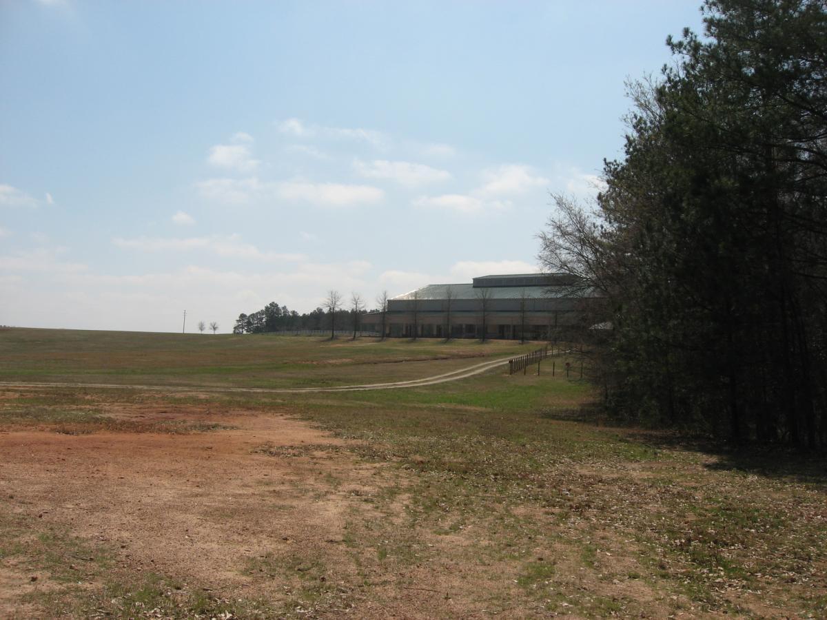 A wide-open field with patches of grass and brown soil leads up to a large building in the background. The sky is partly cloudy, and a line of trees is visible on the right side of the image. A dirt path winds through the landscape, connecting the foreground to the building. Heritage Park mountain bike trail.