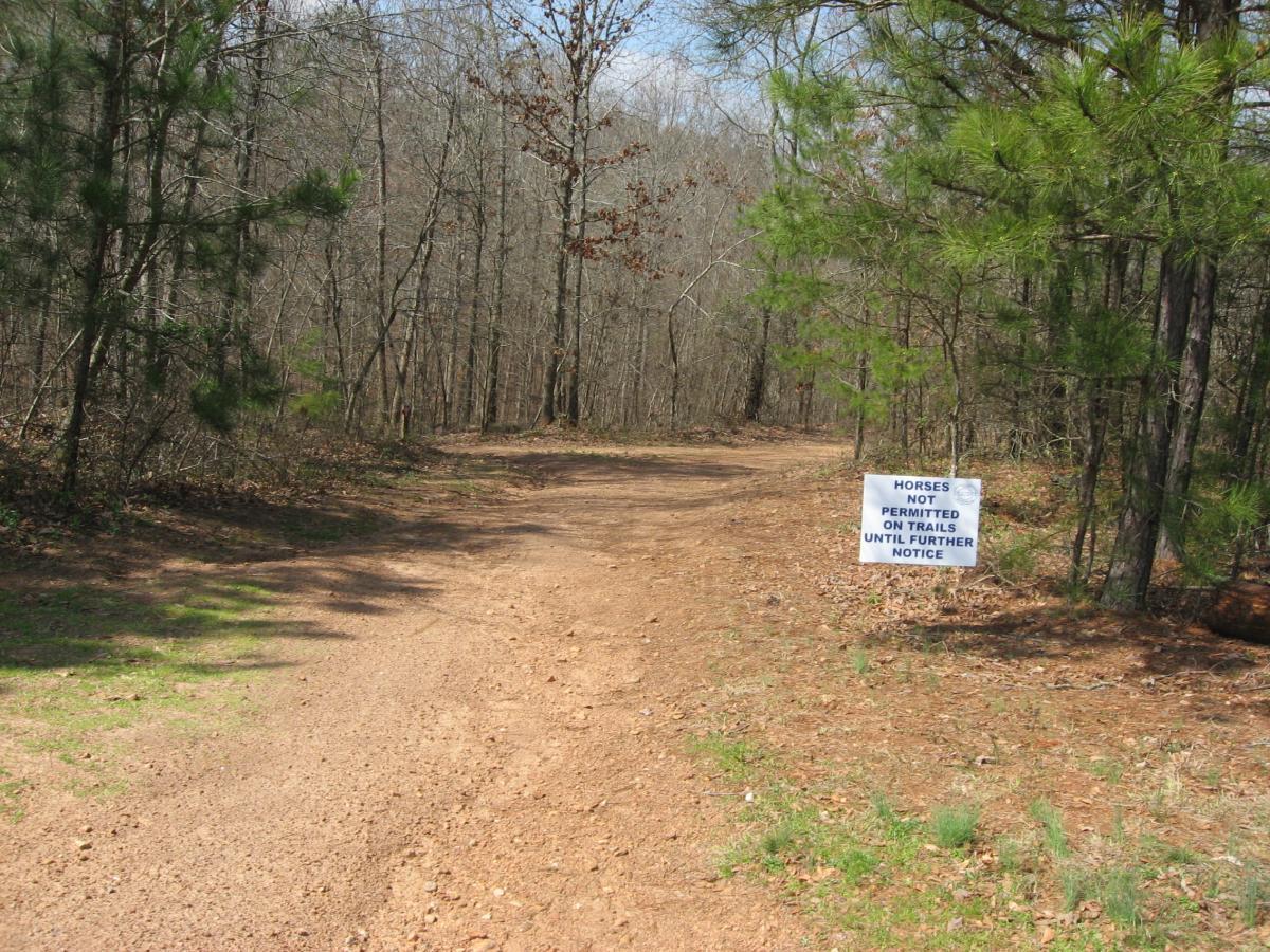 A dirt path surrounded by trees, leading into a wooded area, featuring a white sign that reads, "HORSES NOT PERMITTED ON TRAILS UNTIL FURTHER NOTICE." Heritage Park mountain bike trail.