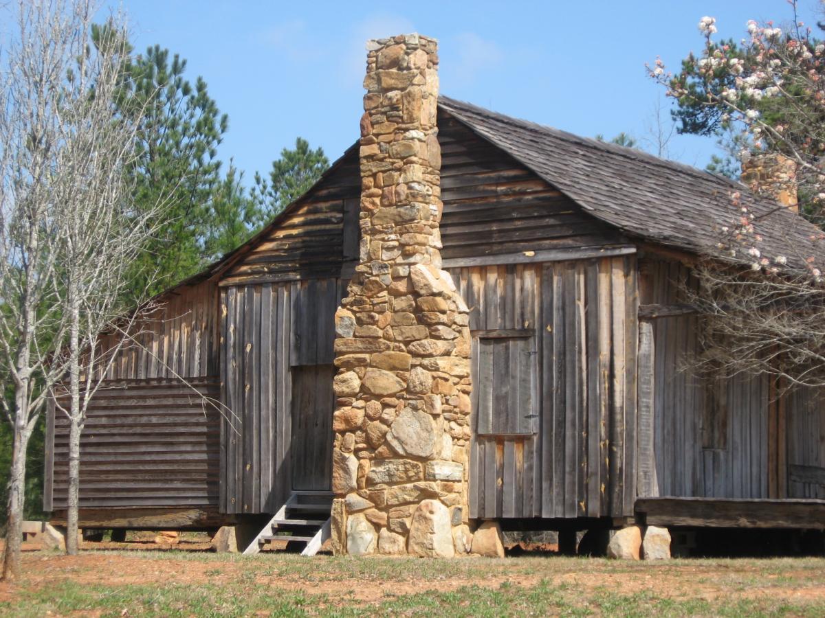 Alt text: A rustic wooden building with a prominent stone chimney, surrounded by trees and grass. The building features weathered wooden siding and a staircase leading to a door on the side, set against a clear blue sky. Heritage Park mountain bike trail.