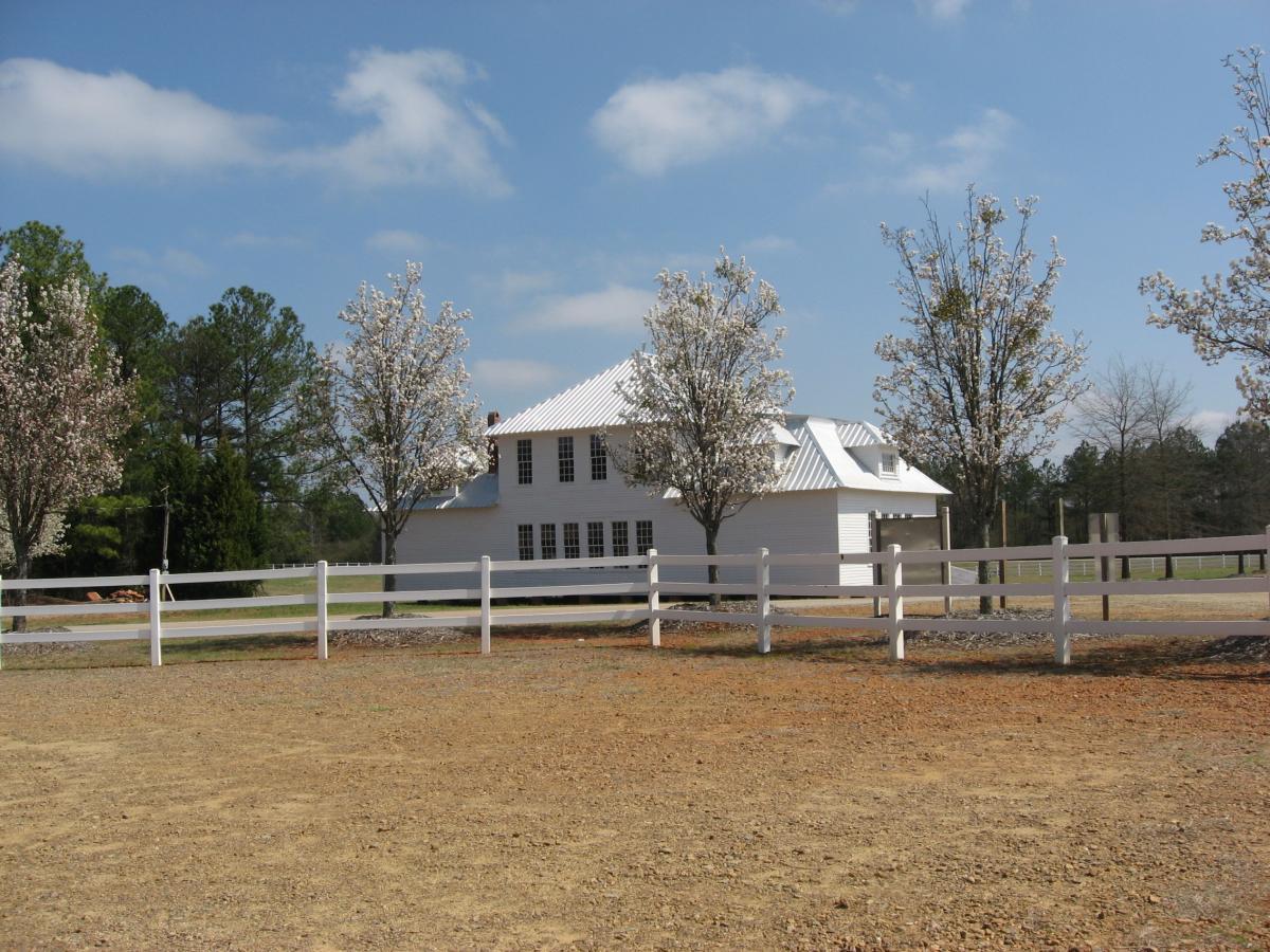 A white building with a metal roof surrounded by blooming trees and a white fence, set against a clear blue sky. The foreground features a dirt area with gravel. Heritage Park mountain bike trail.