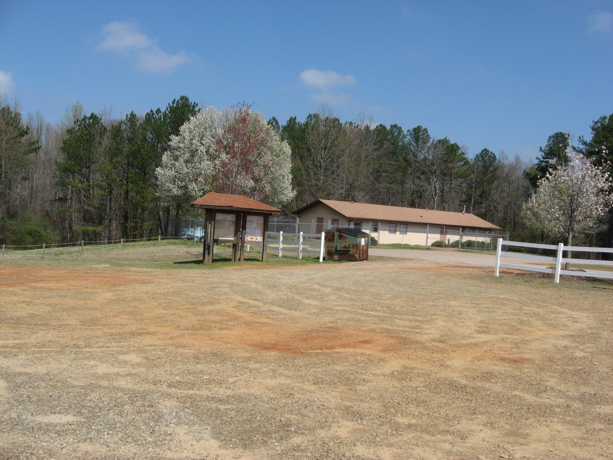 A grassy area with a gravel lot, featuring a small wooden information kiosk under a roof. In the background, a light-colored building is visible, surrounded by trees and a white fence. The sky is partly cloudy, and there are flowering trees nearby. Heritage Park mountain bike trail.