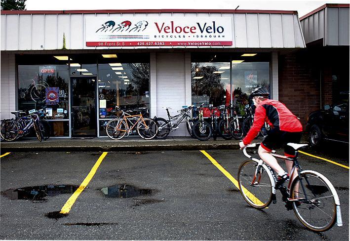 A cyclist in a red jacket rides past the Veloce Velo bicycle shop, which features a variety of bikes displayed outside. The store has an open sign and is set in a parking lot with yellow lines and some puddles.