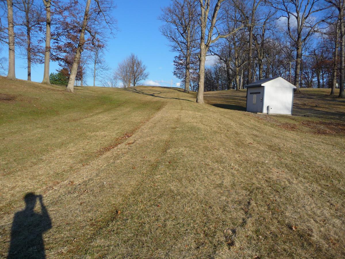 A grassy hillside with bare trees scattered throughout, leading up to a distant rise. A small, white shed is visible to the right, and a person's shadow is cast in the foreground. The sky is clear with a few clouds. Murdock Park Trail mountain bike trail.