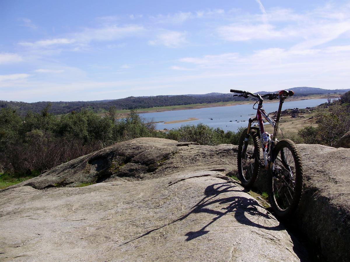 A mountain bike resting on a large rock, overlooking a serene lake and rolling hills under a clear blue sky. The scene is bathed in natural light, highlighting the bike's details and the surrounding greenery. Granite Bay Trail mountain bike trail.