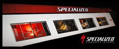 Exterior view of a Specialized Concept Store, featuring large display windows showcasing bicycles and promotional graphics. The store has a red roof and the word "SPECIALIZED" prominently displayed above the entrance.