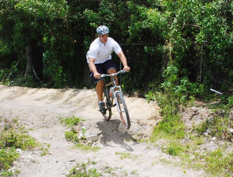 A man wearing a helmet is riding a mountain bike on a dirt path surrounded by greenery. He is lifting the front wheel off the ground, showcasing an action shot while navigating through a trail with a slight incline. Quiet Waters Park mountain bike trail.