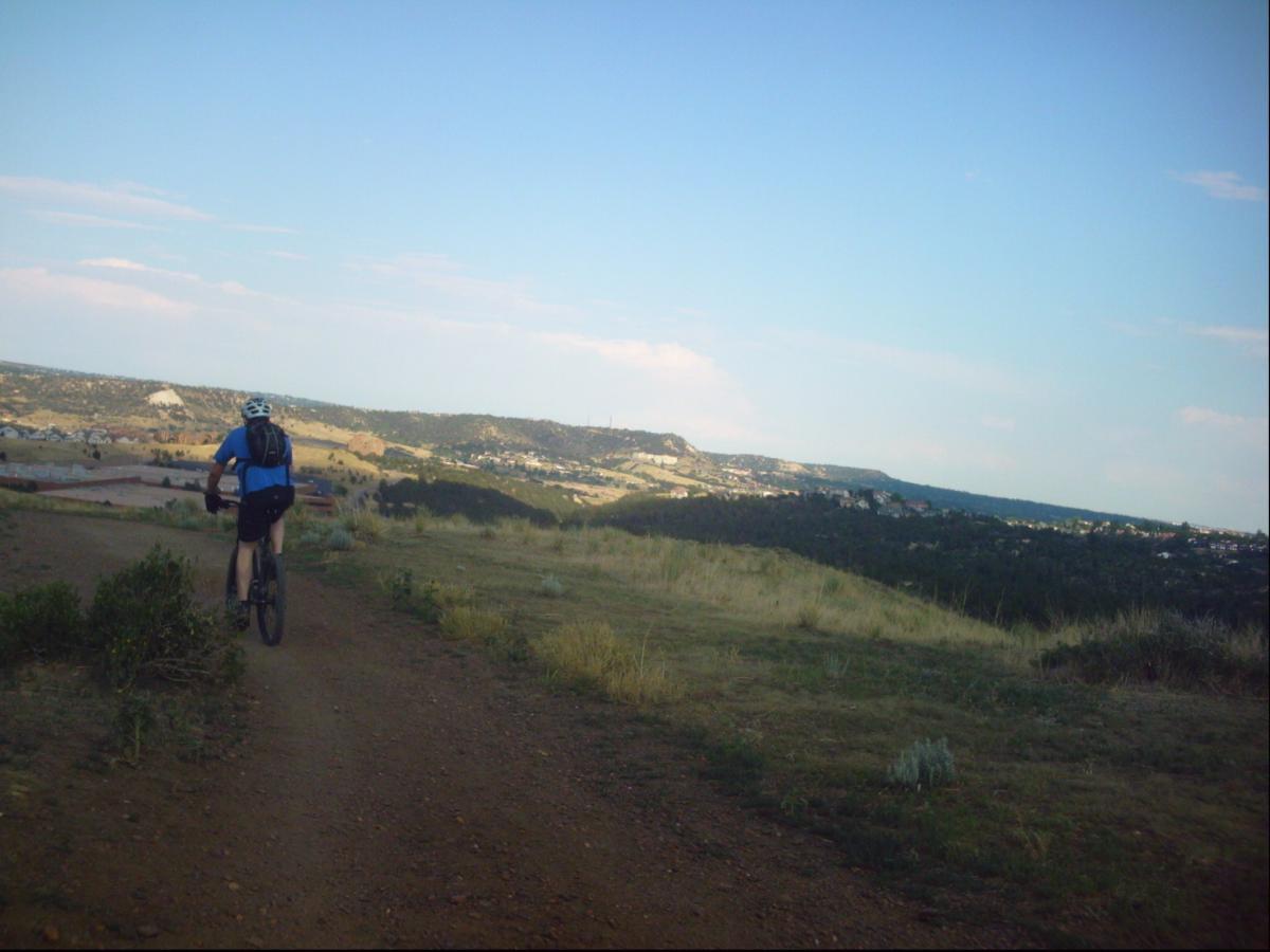 A mountain biker riding on a dirt path surrounded by grassy terrain, with a panoramic view of hills and distant homes under a clear blue sky. Ute Valley Park mountain bike trail.