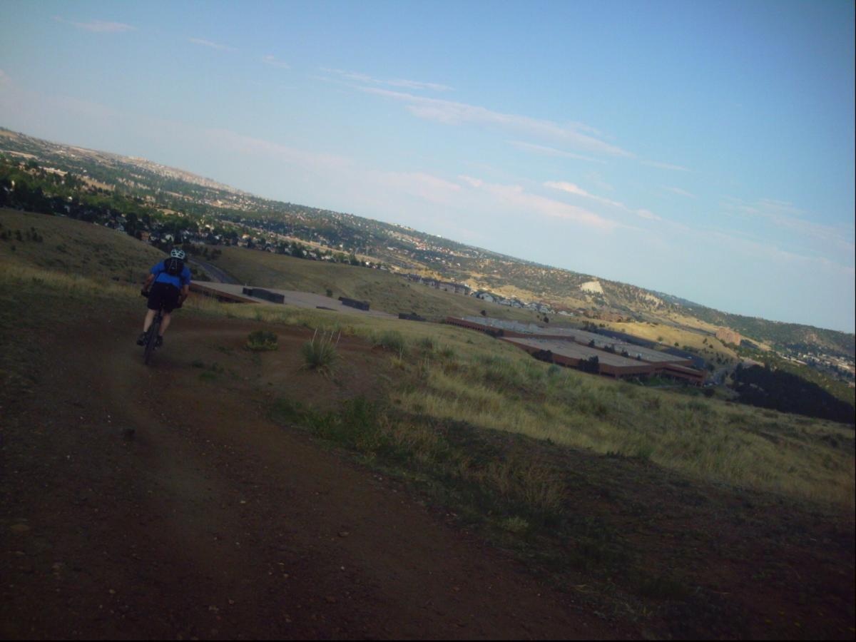 A person riding a mountain bike along a dirt trail on a hillside, with a view of a valley and distant mountains under a clear sky. Ute Valley Park mountain bike trail.