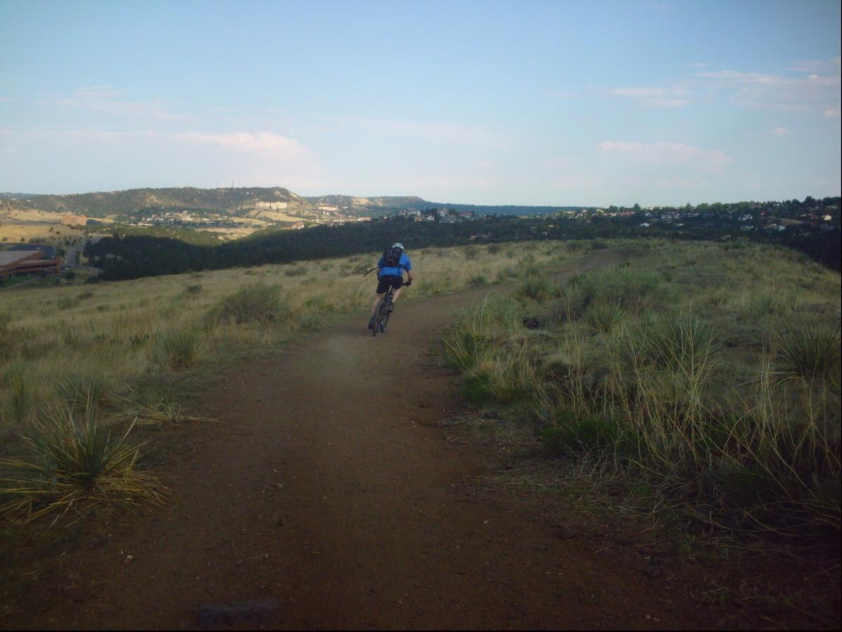 A mountain biker riding on a dirt trail through a grassy landscape, with a view of rolling hills and distant town below under a clear blue sky. Ute Valley Park mountain bike trail.
