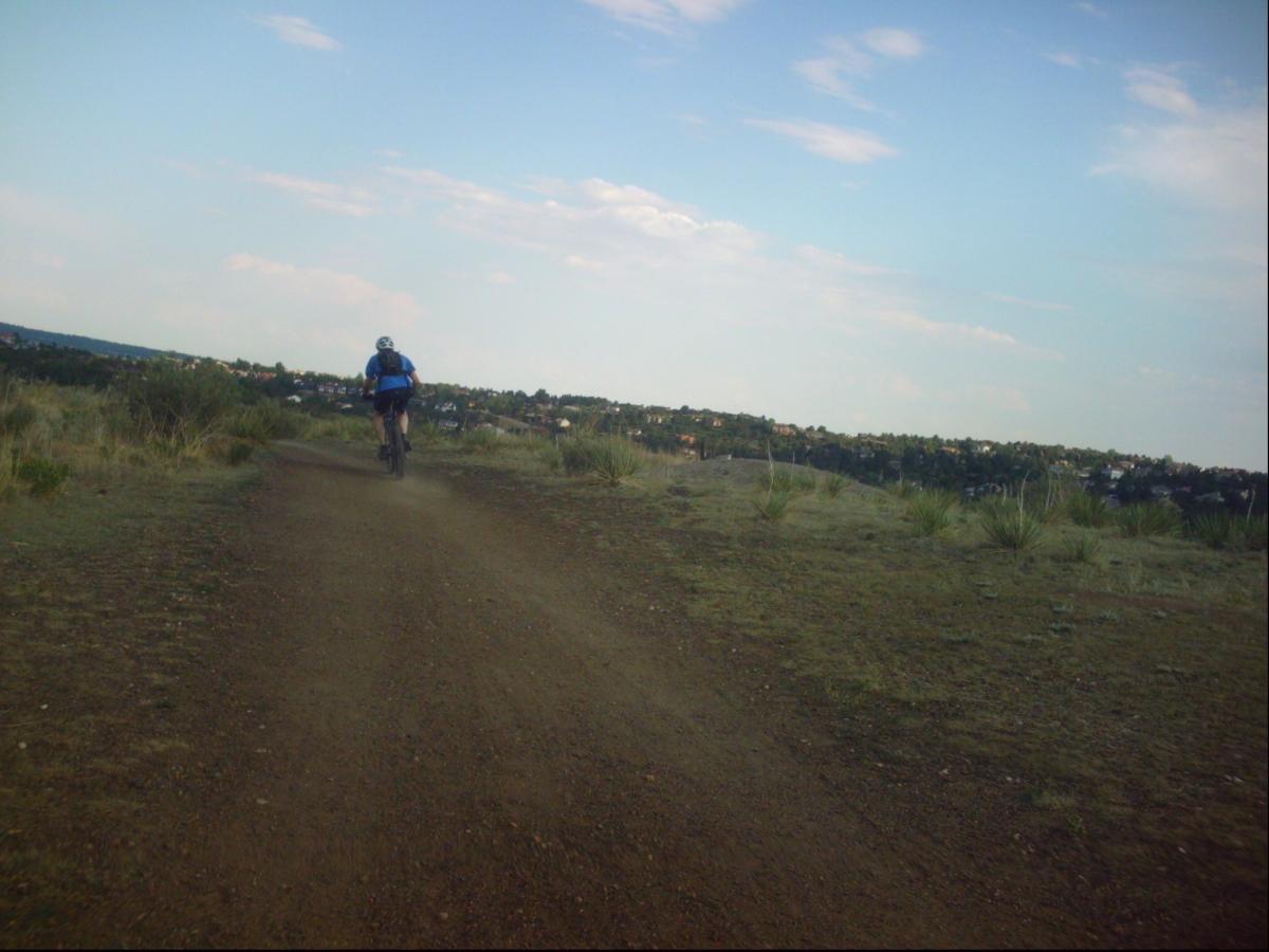 A person riding a mountain bike along a dirt path, with a grassy landscape and distant hills in the background under a partly cloudy sky. Ute Valley Park mountain bike trail.