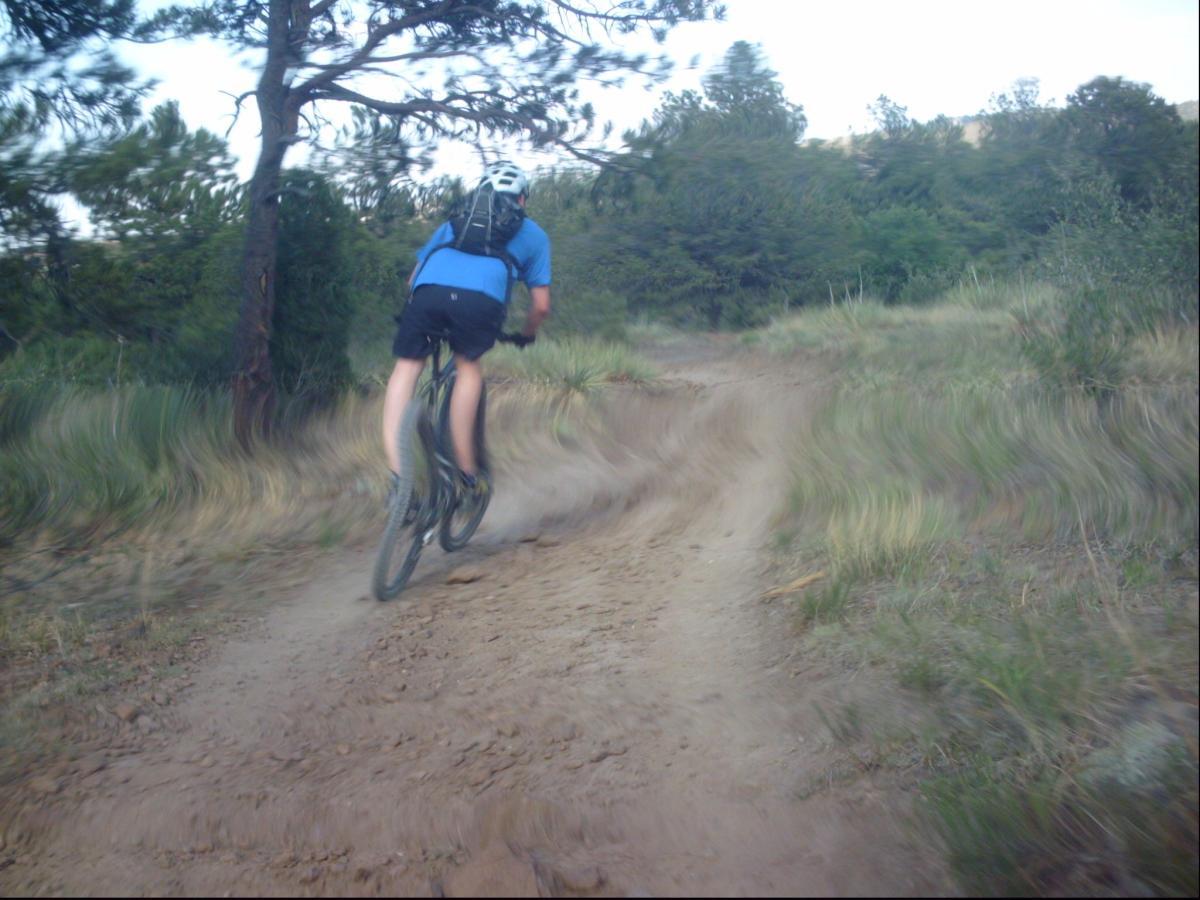A person riding a mountain bike along a dirt trail surrounded by trees and greenery. The cyclist is viewed from behind, wearing a blue shirt and shorts, with a backpack and a helmet. The trail appears winding and uneven, conveying a sense of adventure in a natural outdoor setting. Ute Valley Park mountain bike trail.